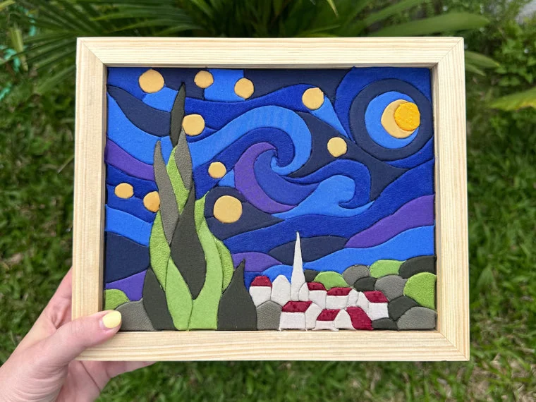 A finished Kinusaiga night sky panel in a pale wood frame, held in one hand outdoors